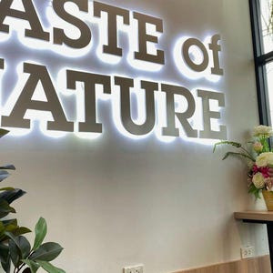 May include: A white wall with a sign that reads "Taste of Nature" in large, silver letters. The sign is lit up with white lights. There are two gray stools in front of the wall, and a wooden counter with a plant on it.