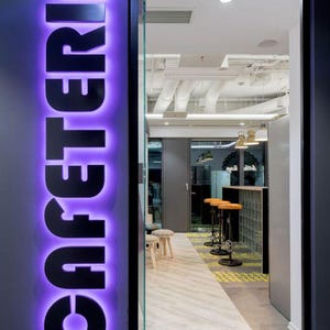 May include: A purple neon sign with the word "CAFETERIA" in black letters. The sign is mounted on a gray wall and is visible through a glass door. The door leads to a room with a white ceiling and a tiled floor.