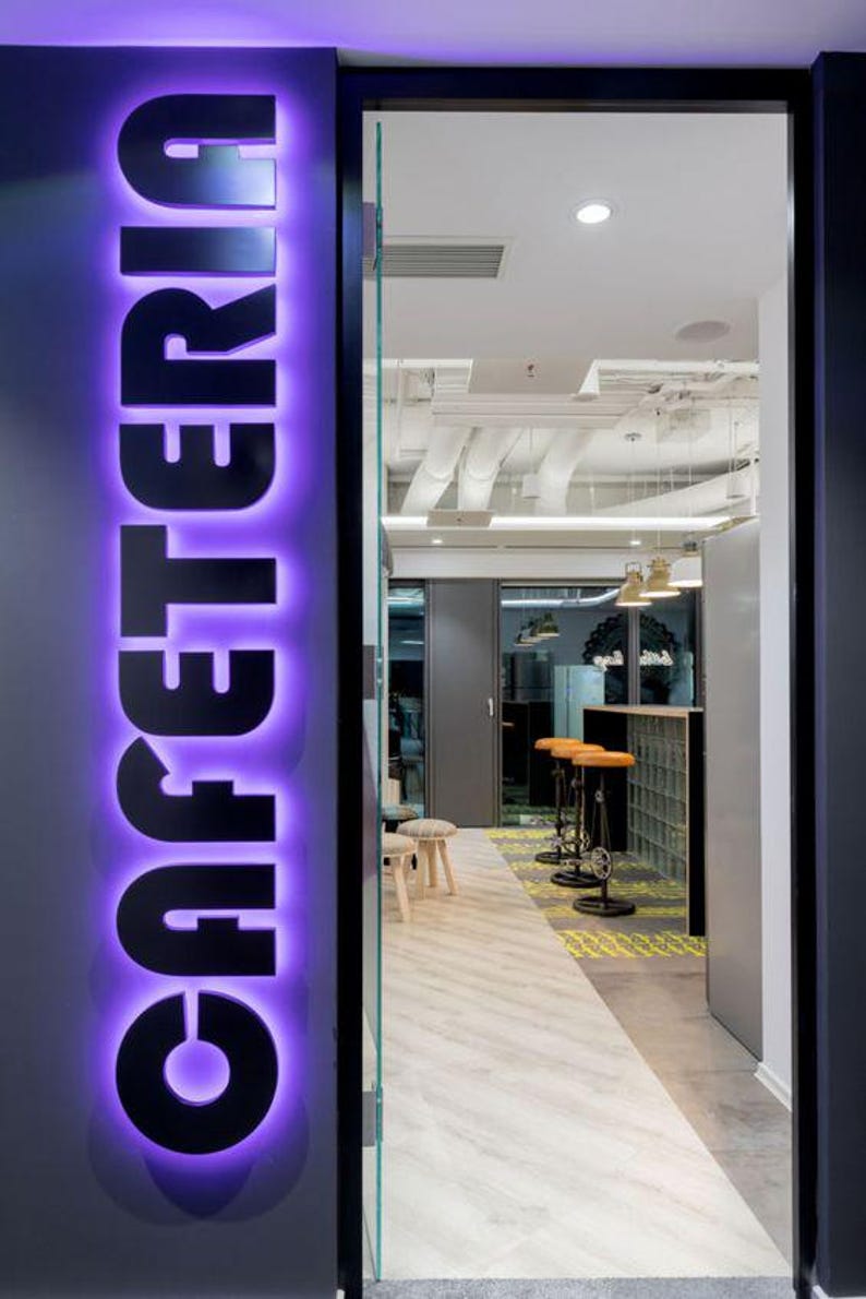 May include: A purple neon sign with the word "CAFETERIA" in black letters. The sign is mounted on a gray wall and is visible through a glass door. The door leads to a room with a white ceiling and a tiled floor.