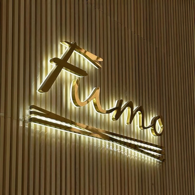 May include: A gold-colored sign with the word "Fumo" illuminated with white lights against a wood-paneled wall.
