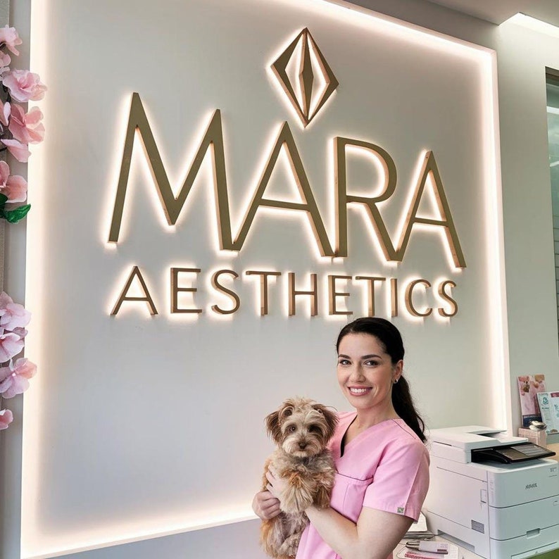 May include: A woman in a pink shirt holds a small brown dog in her arms. The woman is standing in front of a white wall with a gold sign that reads "MARA AESTHETICS".