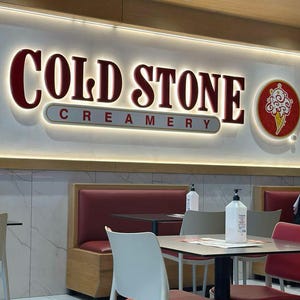 May include: A red and white sign with the text "COLD STONE CREAMERY" in white letters. The sign is illuminated with white lights. There are two tables with chairs in the foreground, and a red leather booth in the background.