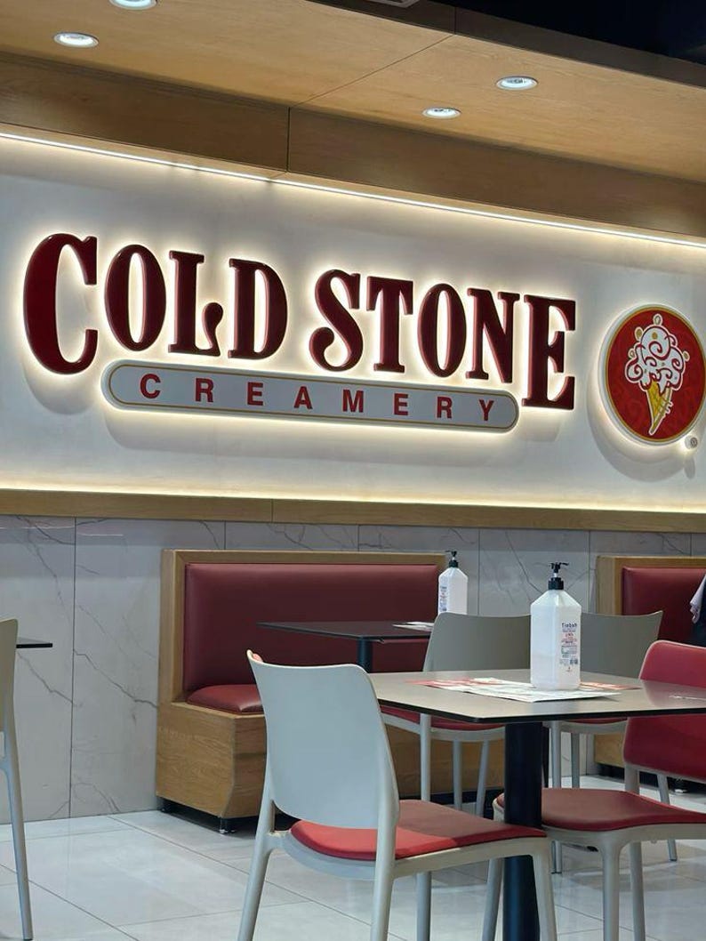 May include: A red and white sign with the text "COLD STONE CREAMERY" in white letters. The sign is illuminated with white lights. There are two tables with chairs in the foreground, and a red leather booth in the background.