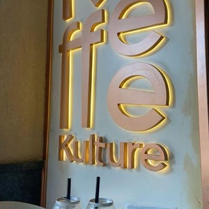 May include: Two iced coffee drinks in clear cans with straws. The cans have the text "Koffee Kultur" printed on them. A black purse with a gold chain is on the table.