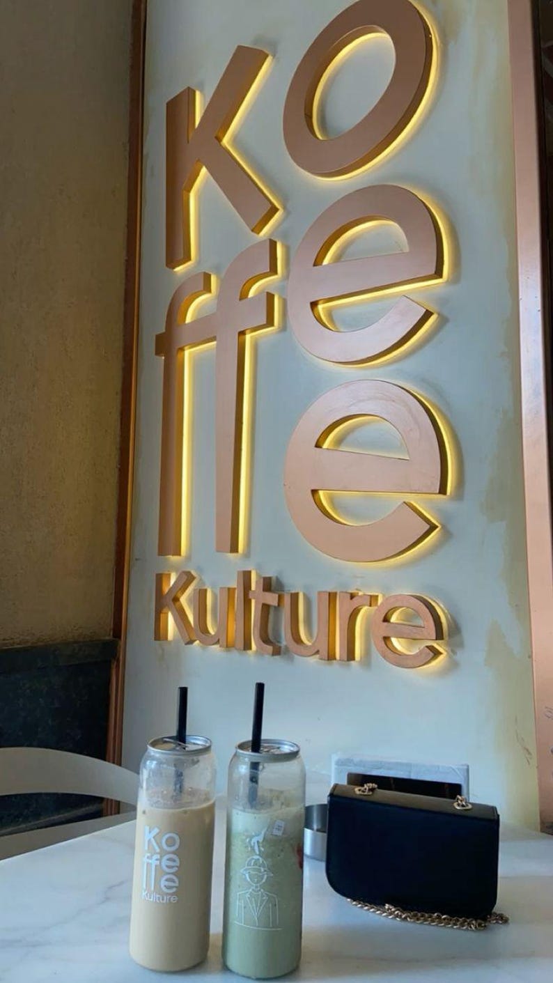 May include: Two iced coffee drinks in clear cans with straws. The cans have the text "Koffee Kultur" printed on them. A black purse with a gold chain is on the table.