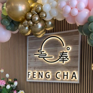 May include: A wooden sign with a white border and a black logo that says "Feng Cha" with a stylized circle and Chinese characters. The sign is decorated with a garland of gold, white, and pink balloons.
