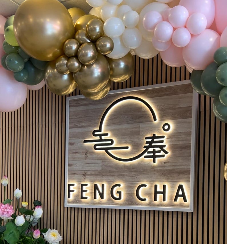 May include: A wooden sign with a white border and a black logo that says "Feng Cha" with a stylized circle and Chinese characters. The sign is decorated with a garland of gold, white, and pink balloons.