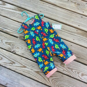 May include: Blue overalls with a dinosaur print. The overalls have red and white gingham trim on the legs.