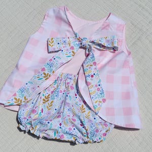 May include: A baby outfit featuring a pink and white gingham top with a large bow in the back. The bow and bloomers have a floral pattern with pink, blue, and yellow accents. The outfit is designed for a baby.