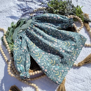 May include: A teal blue baby dress with a floral print featuring small white, pink, and yellow flowers. The dress has long sleeves and a gathered neckline.