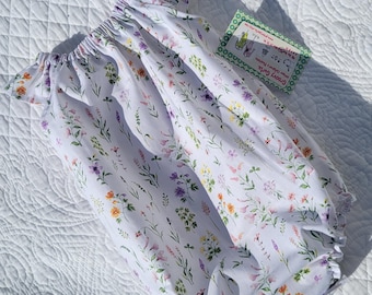 Puede incluir: Un pelele blanco para bebé con estampado floral de pequeñas flores rosas, moradas, amarillas y verdes. El pelele tiene un cuello con volantes y mangas cortas.