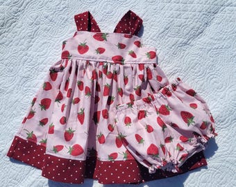 May include: A pink and red strawberry print dress with straps and a matching pair of bloomers. The dress has a burgundy and white polka dot trim.