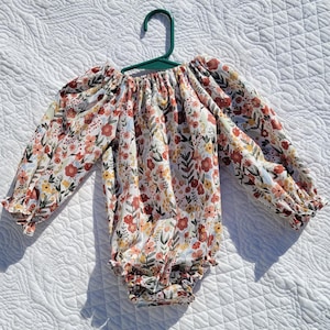 May include: A white long-sleeved baby onesie with a floral print in shades of red, yellow, and blue. The onesie has a gathered neckline and a ruffled hem.
