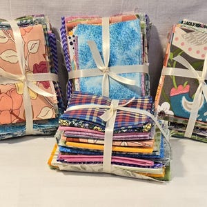 May include: Three bundles of fabric squares tied with ivory ribbon. The fabric features floral, bird, and plaid patterns in various colors. The bundles are stacked, showcasing a variety of designs and textures, perfect for quilting or crafting projects.