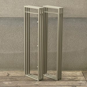 Puede incluir: Dos percheros rectangulares de metal gris claro. Cada perchero tiene una serie de barras verticales y una barra horizontal en la parte superior. Los percheros están colocados sobre una superficie de madera contra una pared gris texturizada.