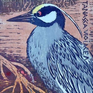 May include: A woodcut print of a bird, featuring a yellow and black head, red eye, and gray body with white accents. The bird is perched on a branch, with text in the background. The artwork has a rustic, vintage aesthetic.