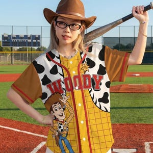 May include: A yellow baseball jersey with a Woody cartoon design. The jersey has brown sleeves and a red trim. The front features the word "WOODY" in red letters and a cartoon image of Woody. The jersey is worn with a brown cowboy hat.