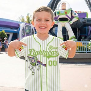 Op de afbeelding: Een wit en groen gestreept honkbalshirt met de groene woorden "Lightyear" en "Buzz", en een afbeelding van Buzz Lightyear. Het shirt heeft het nummer "00". Een kind draagt het shirt.