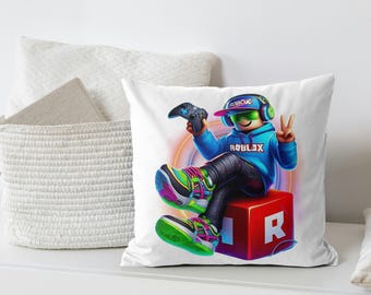 May include: White throw pillow featuring a cartoon gamer character wearing a blue hoodie and holding a game controller. The character is sitting on a red cube with the letter "R" on it. The word "ROBLOX" is visible.