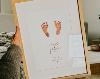 May include: A framed print featuring two copper-colored baby footprints centered at the top. Below the prints, the name Tilda is written in a script font, followed by the date 19.12.2025, 3435g, 51cm, 15.41Uhr, and a small heart icon.