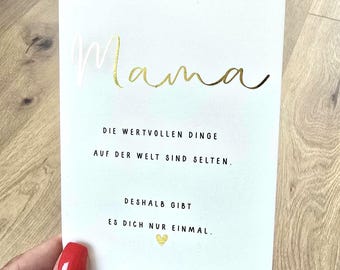 May include: A white card with gold foil lettering that says "Mama". Below the word "Mama" are the words "DIE WERTVOLLEN DINGE AUF DER WELT SIND SELTEN. DESHALB GIBT ES DICH NUR EINMAL." A small gold heart is at the bottom.