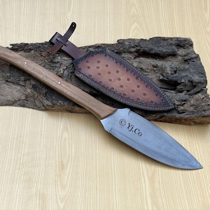 May include: A handcrafted spear with a wooden handle and a metal blade, displayed on a piece of wood. The spear is shown with a leather sheath featuring decorative stitching. The text "© Yj.Co" is printed on the blade.