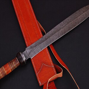 May include: A Damascus steel sword with a patterned blade, wooden handle, and a red leather sheath. The sword has a long, pointed blade and a detailed handle with a rounded pommel. The sheath is stitched and has a strap.