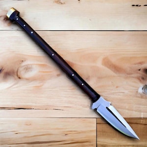 May include: A dark wooden spear with a polished metal blade. The spear has a dark brown shaft with white dot accents and a gold-coloured metal cap on the top. The blade is silver and has a sharp point.