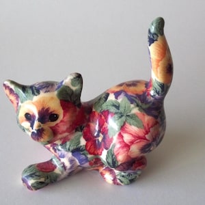 May include: A ceramic cat figurine with a floral pattern in shades of red, purple, yellow, and green. The cat is in a playful pose with its tail up. The figurine is a decorative item.