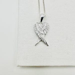 Puede incluir: Un collar con colgante plateado con un dise&ntilde;o detallado de alas. El colgante cuelga de una fina cadena. Las alas est&aacute;n intrincadamente detalladas, con cada pluma claramente definida. El collar se muestra sobre un fondo blanco.