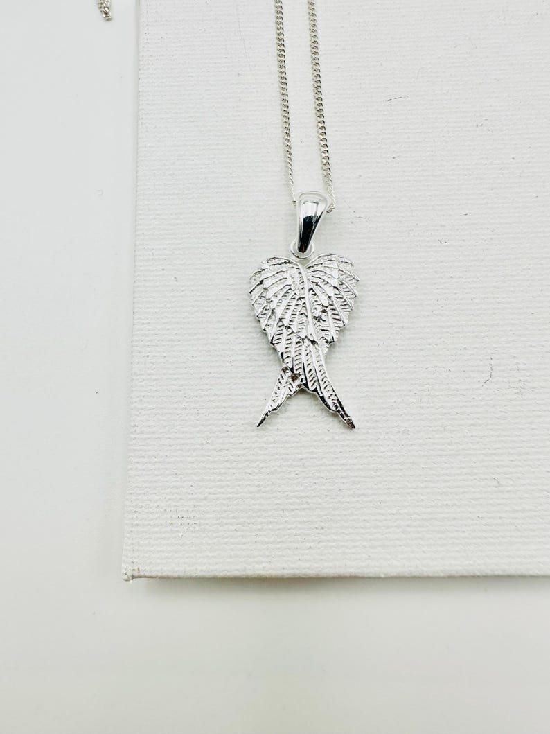 Puede incluir: Un collar con colgante plateado con un dise&ntilde;o detallado de alas. El colgante cuelga de una fina cadena. Las alas est&aacute;n intrincadamente detalladas, con cada pluma claramente definida. El collar se muestra sobre un fondo blanco.