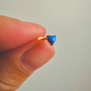 May include: A gold-colored stud earring with a heart-shaped, blue gemstone. The earring is small and delicate, with the gemstone set in a gold bezel. The earring is held between two fingers against a white background.
