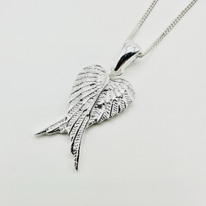 Puede incluir: Un collar con colgante plateado con un dise&ntilde;o de alas detallado. El colgante, suspendido de una delicada cadena, presenta alas intrincadamente detalladas, con cada pluma claramente definida. El collar se muestra sobre un fondo blanco.