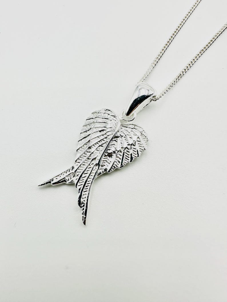 Puede incluir: Un collar con colgante plateado con un dise&ntilde;o de alas detallado. El colgante, suspendido de una delicada cadena, presenta alas intrincadamente detalladas, con cada pluma claramente definida. El collar se muestra sobre un fondo blanco.