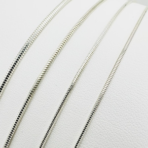 May include: Three silver-coloured snake chains arranged in parallel lines. Each chain features a smooth, flexible, and tightly linked metallic construction with a reflective surface, showcasing a uniform round profile and a consistent polished finish.