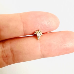Puede incluir: Un pequeño pendiente dorado con un diseño geométrico. El pendiente presenta una piedra central en forma de diamante y un diseño en forma de sol. El pendiente se muestra en un dedo sobre un fondo blanco.