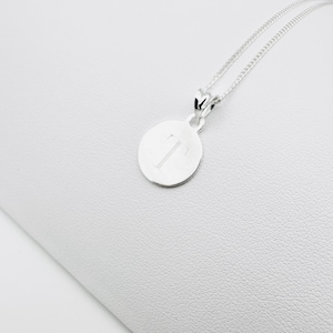 Puede incluir: Un collar con colgante de plata con un dije circular grabado con la letra "T". El colgante cuelga de una fina cadena de plata. La joya se exhibe sobre un fondo blanco, ofreciendo una estética limpia y elegante.