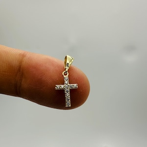 May include: A small, gold-colored cross pendant with embedded clear gemstones. The cross is held between two fingers against a neutral background. The pendant is a religious symbol.