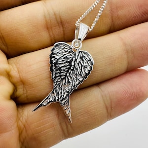 Puede incluir: Un collar con colgante plateado con un dise&ntilde;o de ala detallado. El colgante tiene una superficie texturizada, con intrincados detalles de plumas. La cadena del collar tambi&eacute;n es plateada y el colgante est&aacute; suspendido de un peque&ntilde;o lazo.