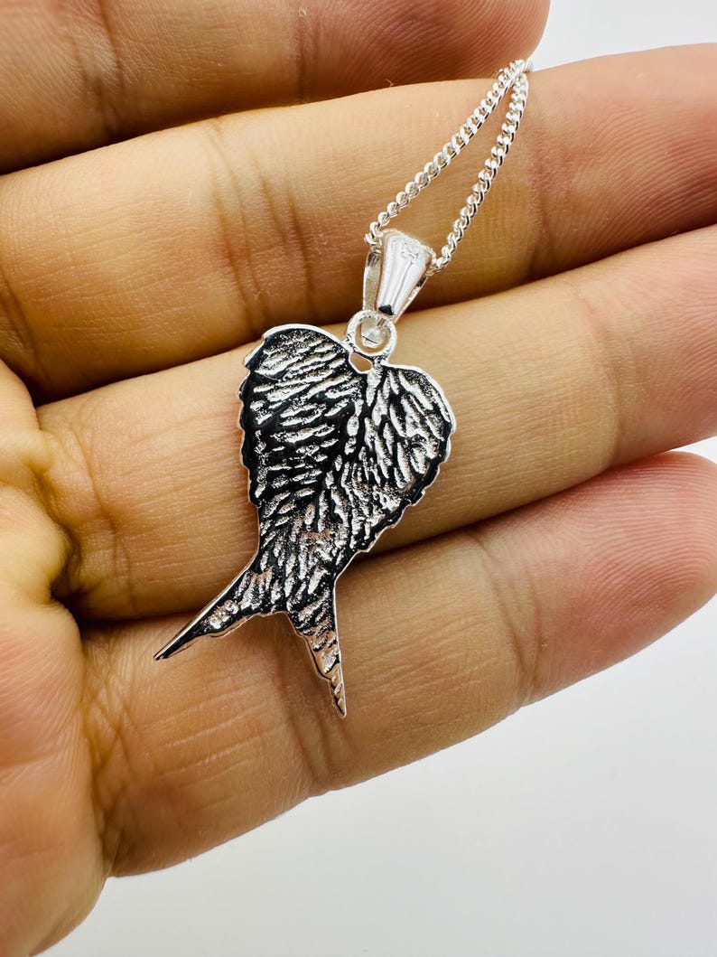 Puede incluir: Un collar con colgante plateado con un dise&ntilde;o de ala detallado. El colgante tiene una superficie texturizada, con intrincados detalles de plumas. La cadena del collar tambi&eacute;n es plateada y el colgante est&aacute; suspendido de un peque&ntilde;o lazo.