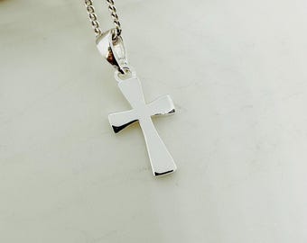 May include: A silver-colored cross pendant on a chain. The cross is a simple, polished design with rounded edges. The chain is a delicate, linked style, and the pendant hangs against a light-colored background.