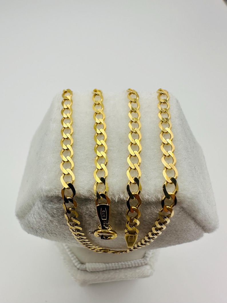 May include: A gold-colored curb chain necklace displayed on a white surface. The necklace is made of interlocking links and features a clasp. The chain is approximately 18 inches long.