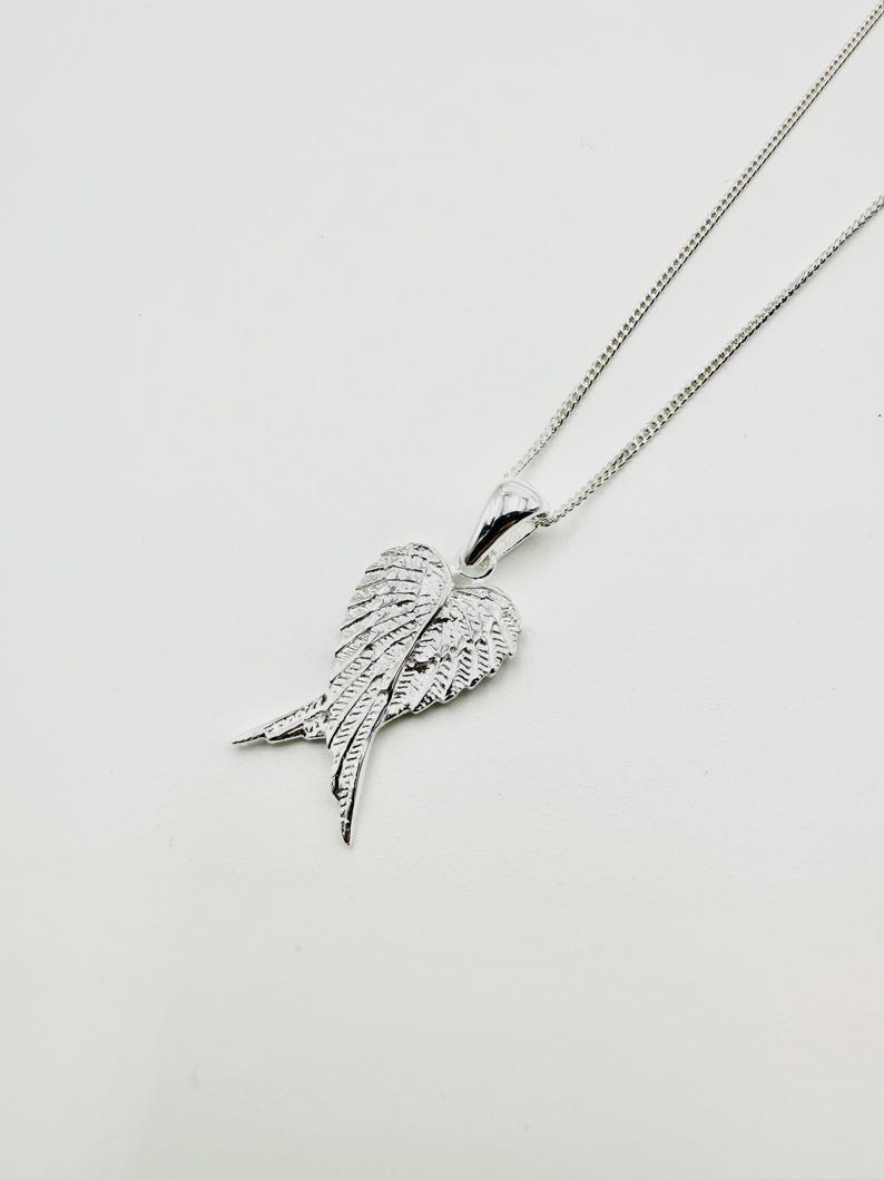 Puede incluir: Un collar plateado con un colgante en forma de alas de &aacute;ngel. Las alas, con texturas detalladas de plumas, cuelgan de una fina cadena. El colgante mide aproximadamente 2,5 cm de ancho.