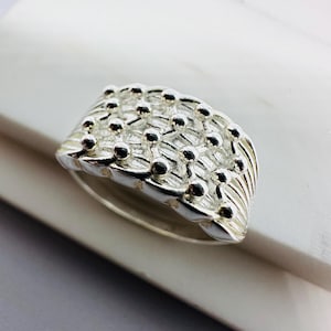 May include: A silver ring with a textured design. The ring features a series of small, raised spheres and curved lines, creating a unique pattern. The ring is set against a neutral background, highlighting its metallic shine.