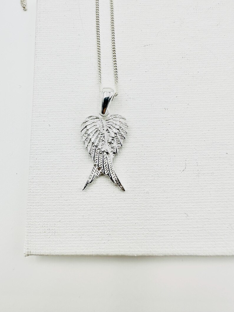 Puede incluir: Un collar plateado con un colgante con dise&ntilde;o de alas de &aacute;ngel. El colgante, suspendido de una fina cadena, presenta detalles de plumas. El collar se exhibe sobre una superficie blanca. Las alas del colgante est&aacute;n intrincadamente elaboradas.