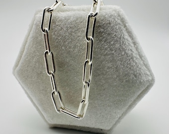 May include: A silver chain necklace with large, rectangular links. The chain is displayed on a light grey, hexagonal, plush surface. The links are evenly spaced and reflect light, creating a bright, shiny appearance. The necklace is a simple, yet elegant piece of jewellery.