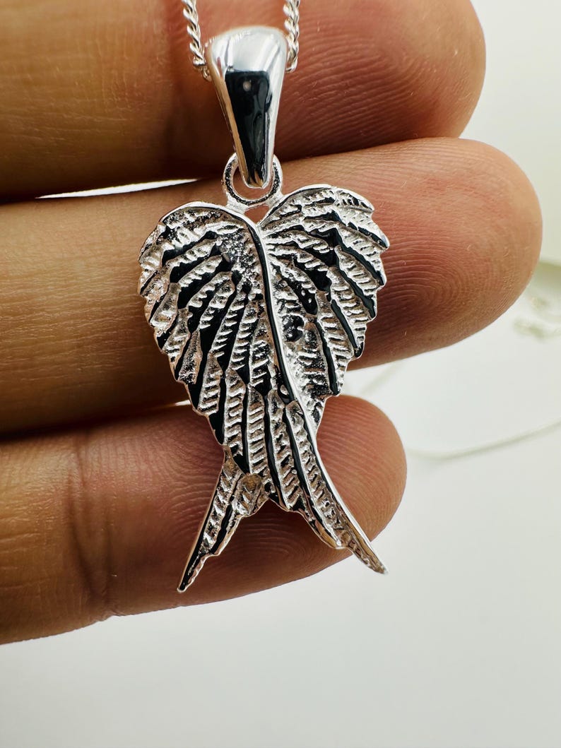 Puede incluir: Un collar con colgante plateado con un dise&ntilde;o de alas detalladas. El colgante tiene una superficie texturizada con intrincados detalles de plumas. La cadena del collar tambi&eacute;n es plateada y parece delicada.