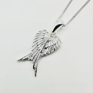 Puede incluir: Collar con colgante plateado en forma de ala. El colgante, con detalles de plumas, cuelga de una fina cadena. El collar se presenta sobre un fondo blanco. El dise&ntilde;o de ala es una caracter&iacute;stica destacada de la joya.