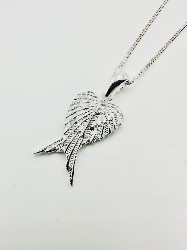 Puede incluir: Collar con colgante plateado en forma de ala. El colgante, con detalles de plumas, cuelga de una fina cadena. El collar se presenta sobre un fondo blanco. El dise&ntilde;o de ala es una caracter&iacute;stica destacada de la joya.