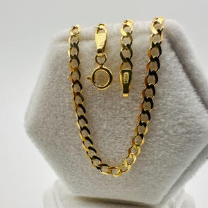 May include: A gold-colored curb chain necklace with a lobster clasp and a separate chain segment with a spring ring clasp. The chain is displayed on a white, textured surface. The links are flat and oval.